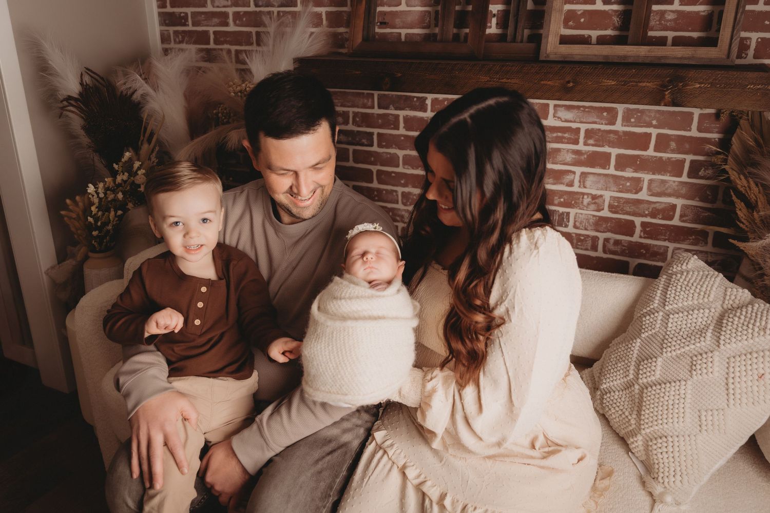 Happy family of four with newborn baby sitting together on couch near brick wall.
