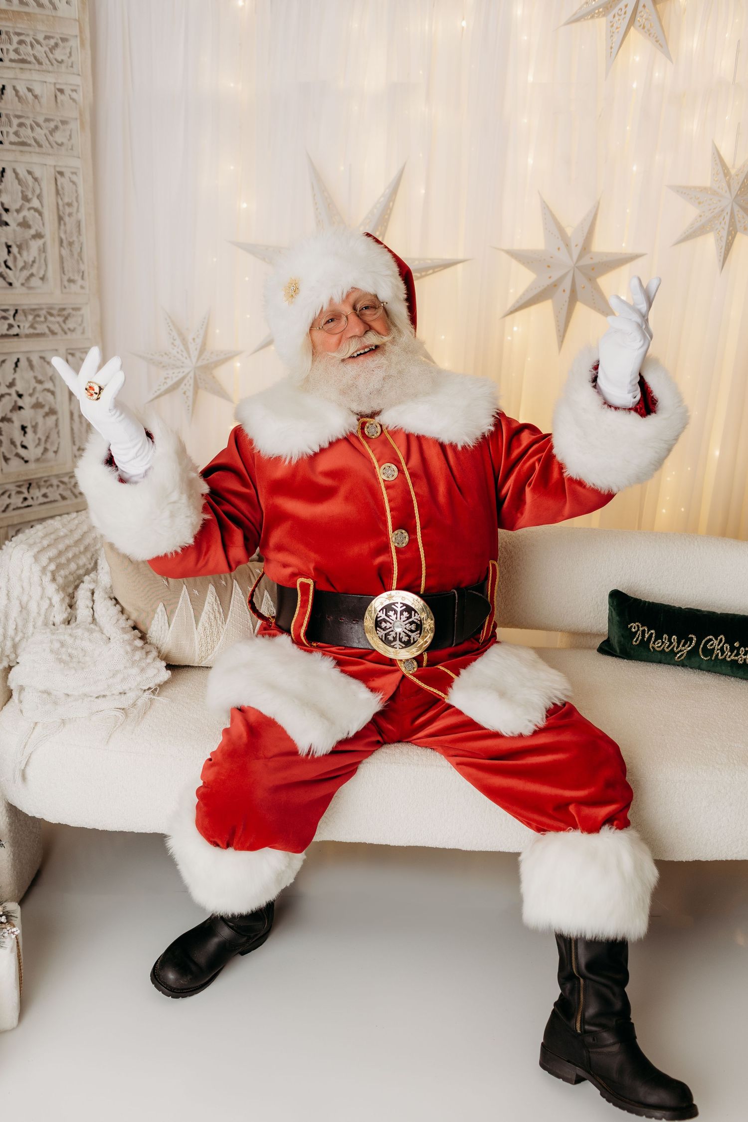 Santa Claus in red suit sits on white sofa, gesturing with gloved hands among star decorations and Christmas lights near Philadelphia.