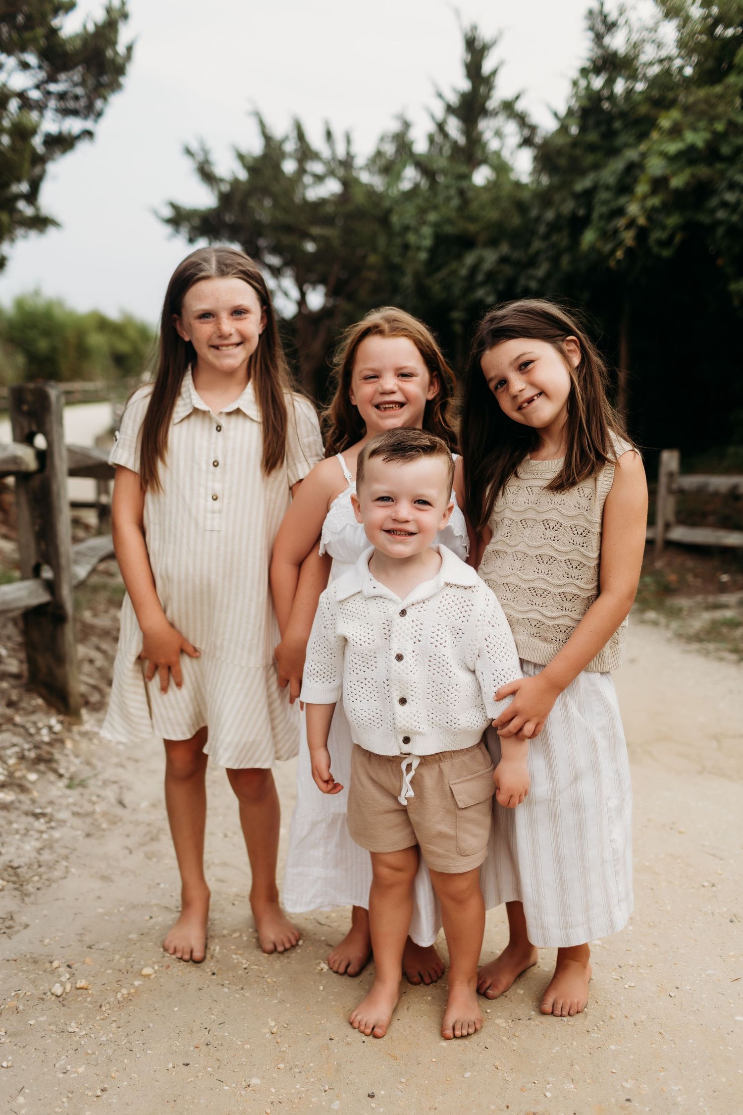 Four barefoot children pose together on a sandy path, dressed in neutral tones, smiling for a family beach photo.