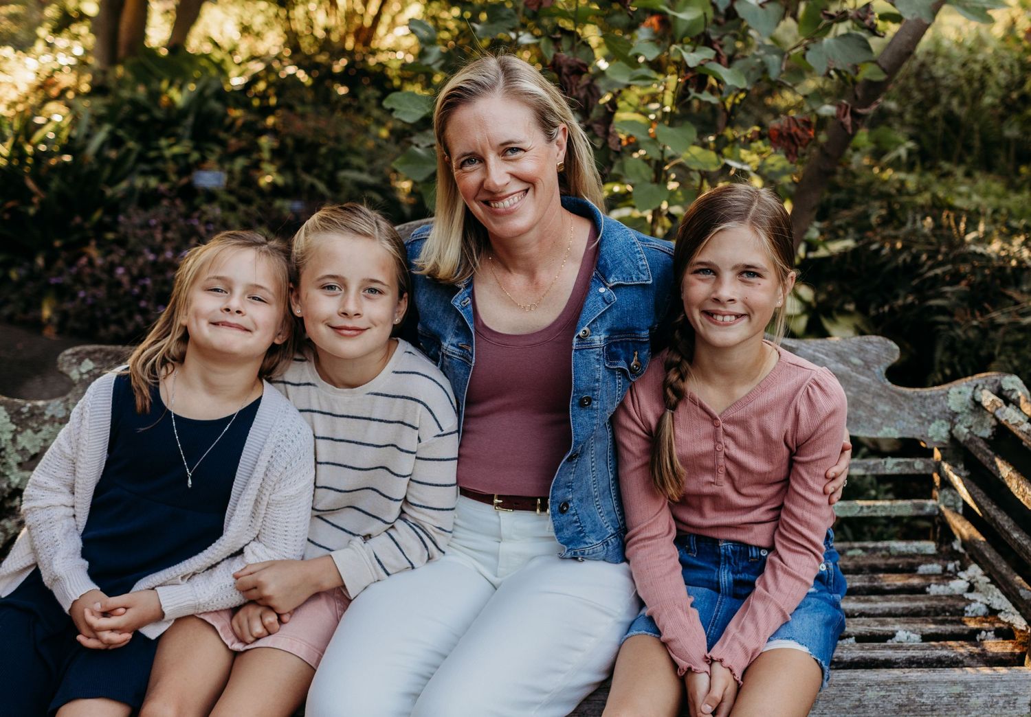 Blonde mother sitting with three daughters on stone bench in lush garden setting.