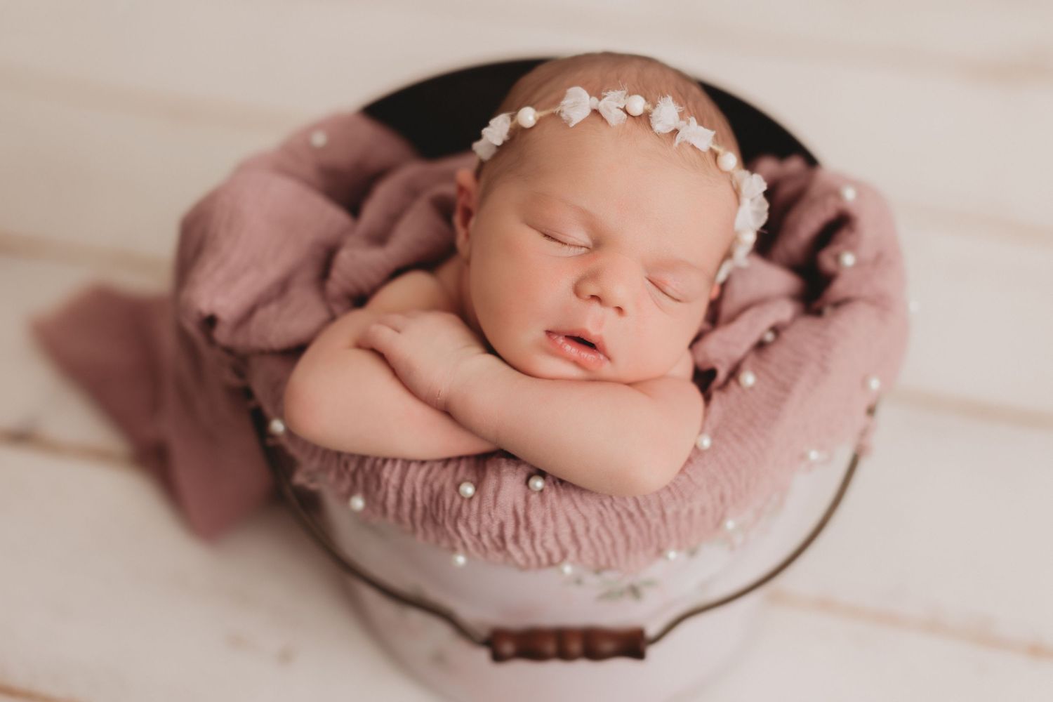Sweet sleeping newborn posed in pink bowl wearing delicate pearl headband on soft fabric backdrop.