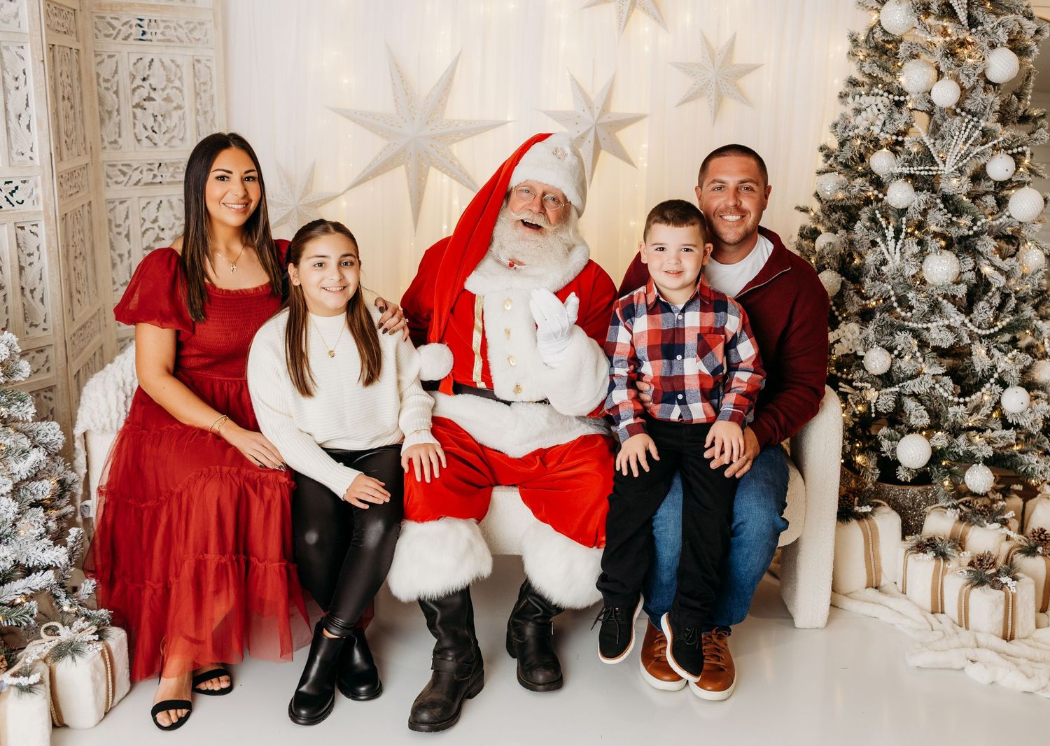 A family of four sits with Santa Claus on a white sofa beside a silver and white decorated Christmas tree.