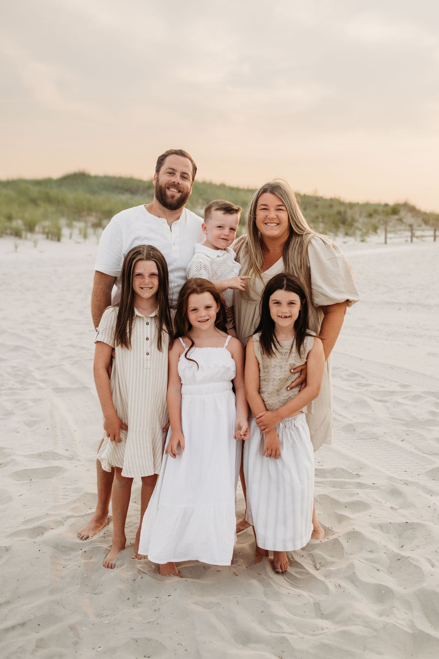 Family of six posing together on a sandy beach at sunset, wearing coordinating neutral and white outfits.