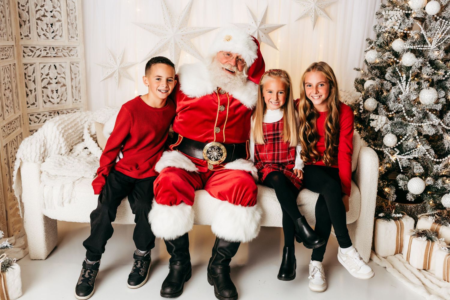 Three children pose with Santa Claus on a white couch beside a decorated Christmas tree in a festive holiday portrait.