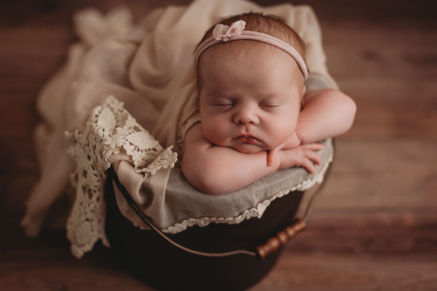 Sleeping newborn baby with pink bow headband posed in dark wooden bucket with lace.