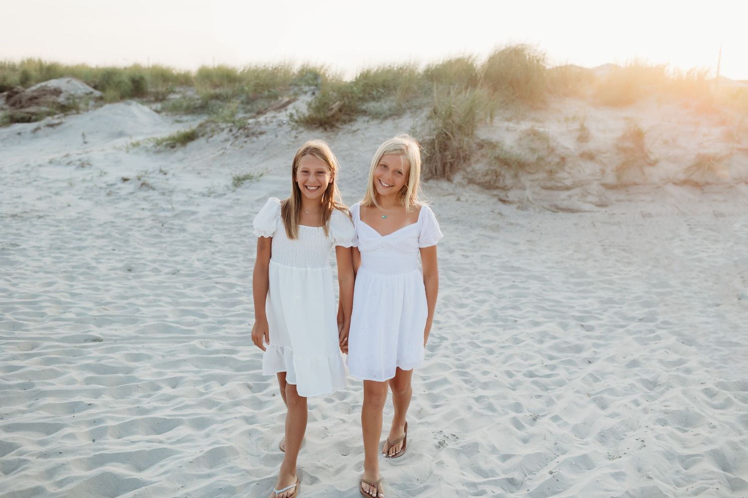 Two people in white dresses walking together on a sandy beach at sunset.