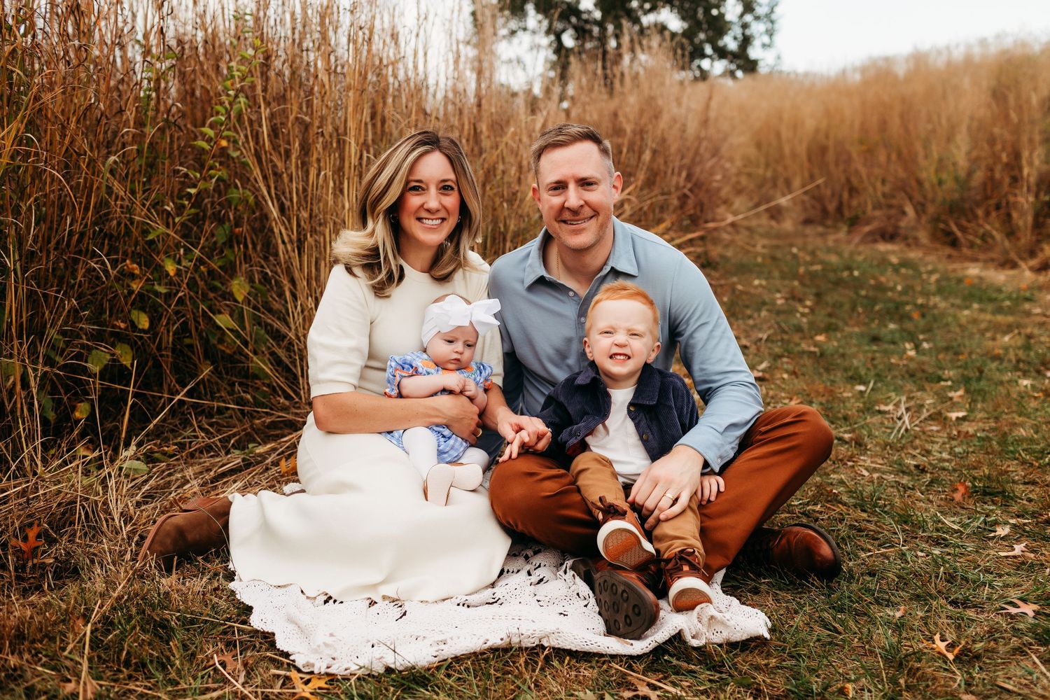 Family of four sitting outdoors in autumn field, parents smiling with baby and toddler on blanket.