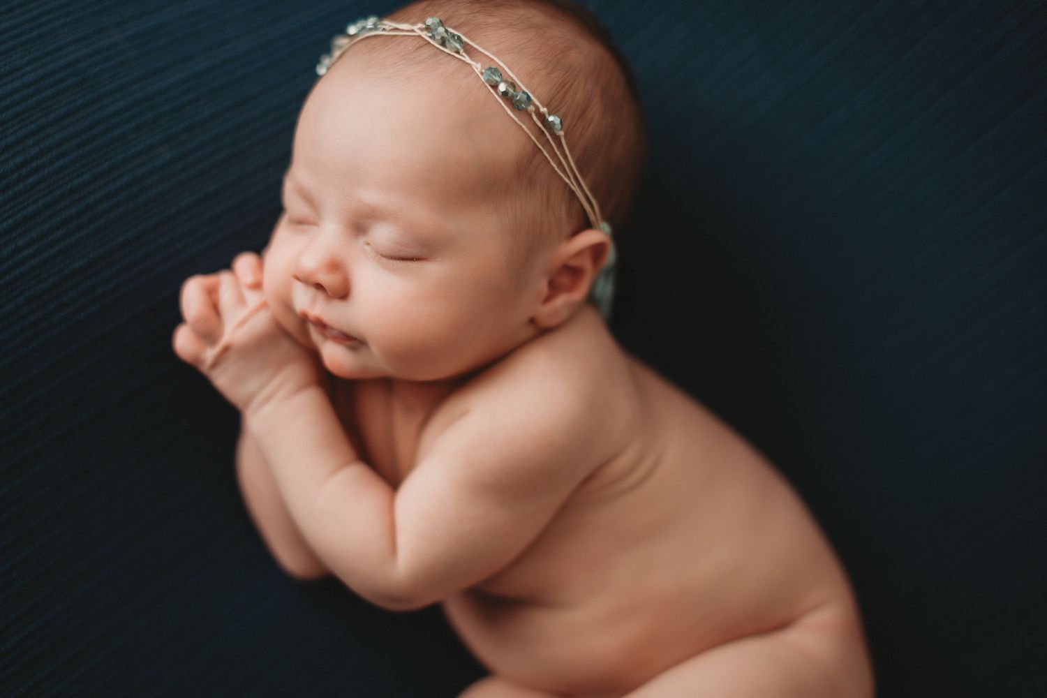 Peaceful newborn baby wearing jeweled headband sleeping on dark blue backdrop with hands near face.