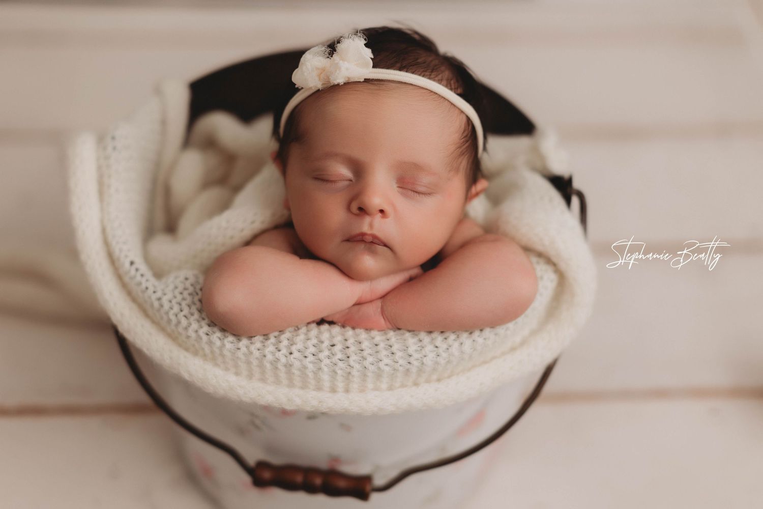 A sleeping newborn wearing a white headband rests peacefully in a cozy knit basket on neutral toned fabric.