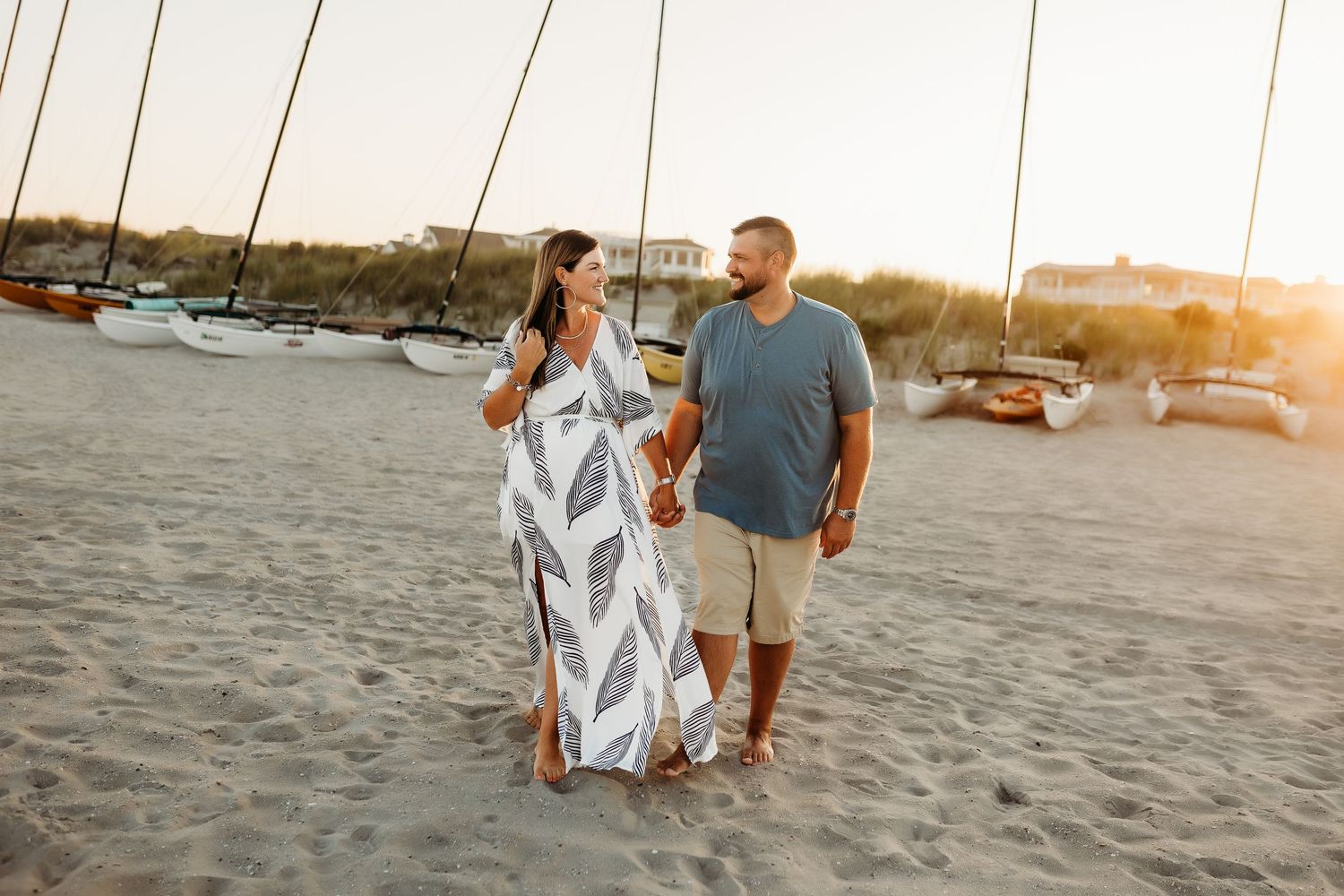 A couple walks hand in hand on a sandy beach at golden hour near sailboats.