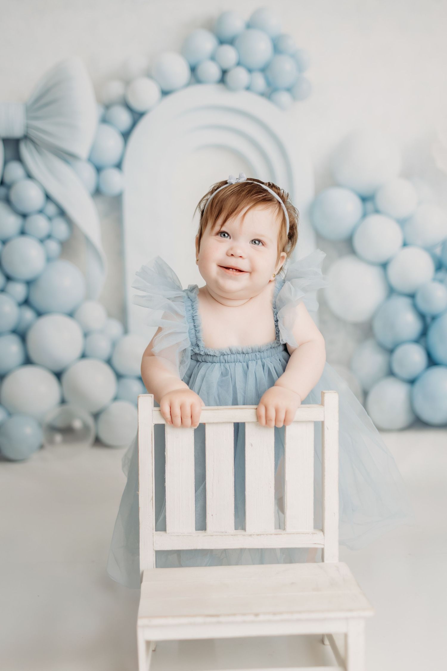 Happy baby girl in blue tulle dress standing at white chair with blue balloon decorations behind her.