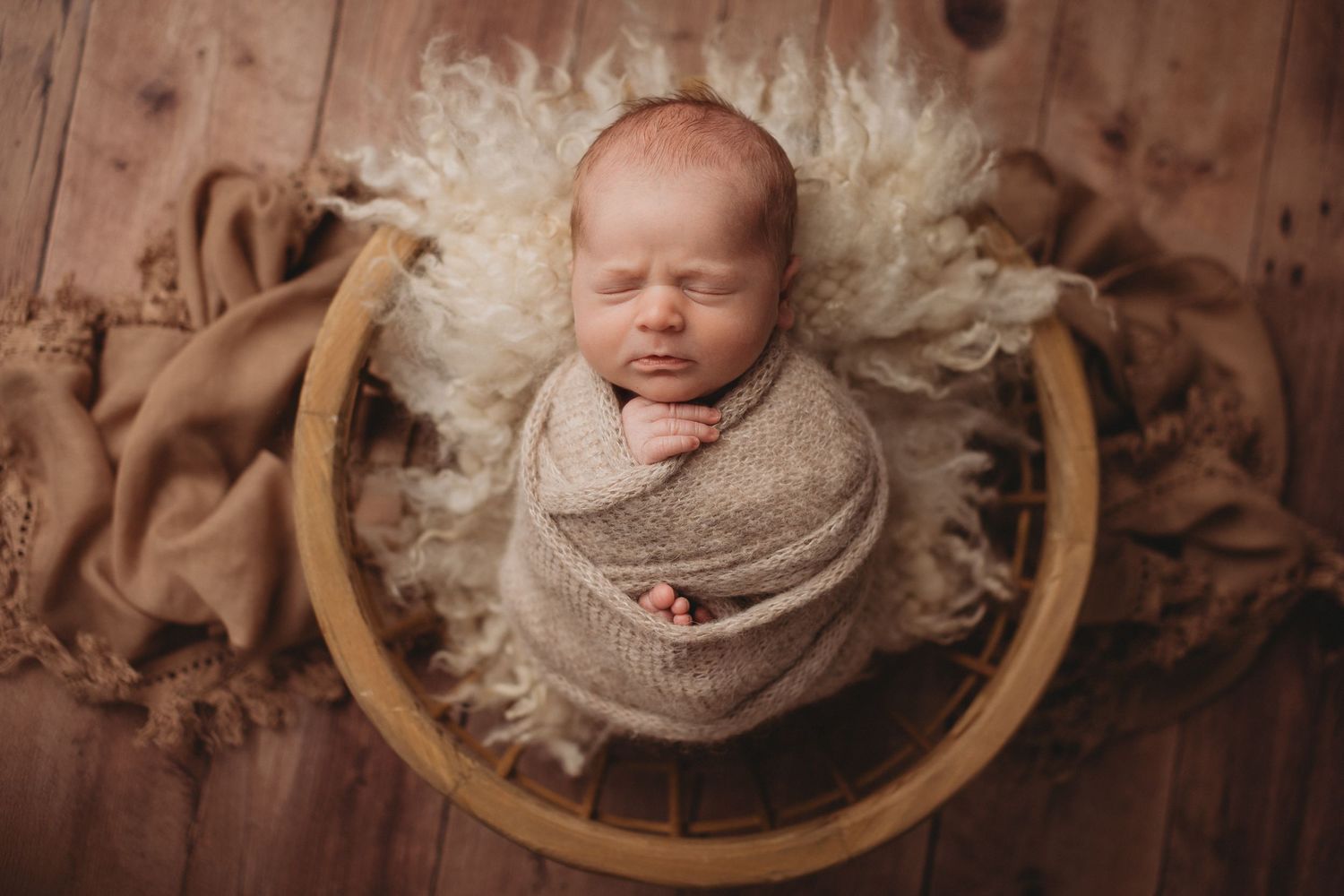 Newborn baby swaddled in beige wrap sleeping in wooden bowl with cream fur lining.
