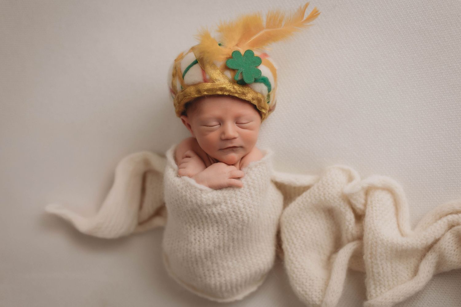 A newborn baby wearing a felt crown with a feather lies peacefully swaddled in a cozy white knit blanket.