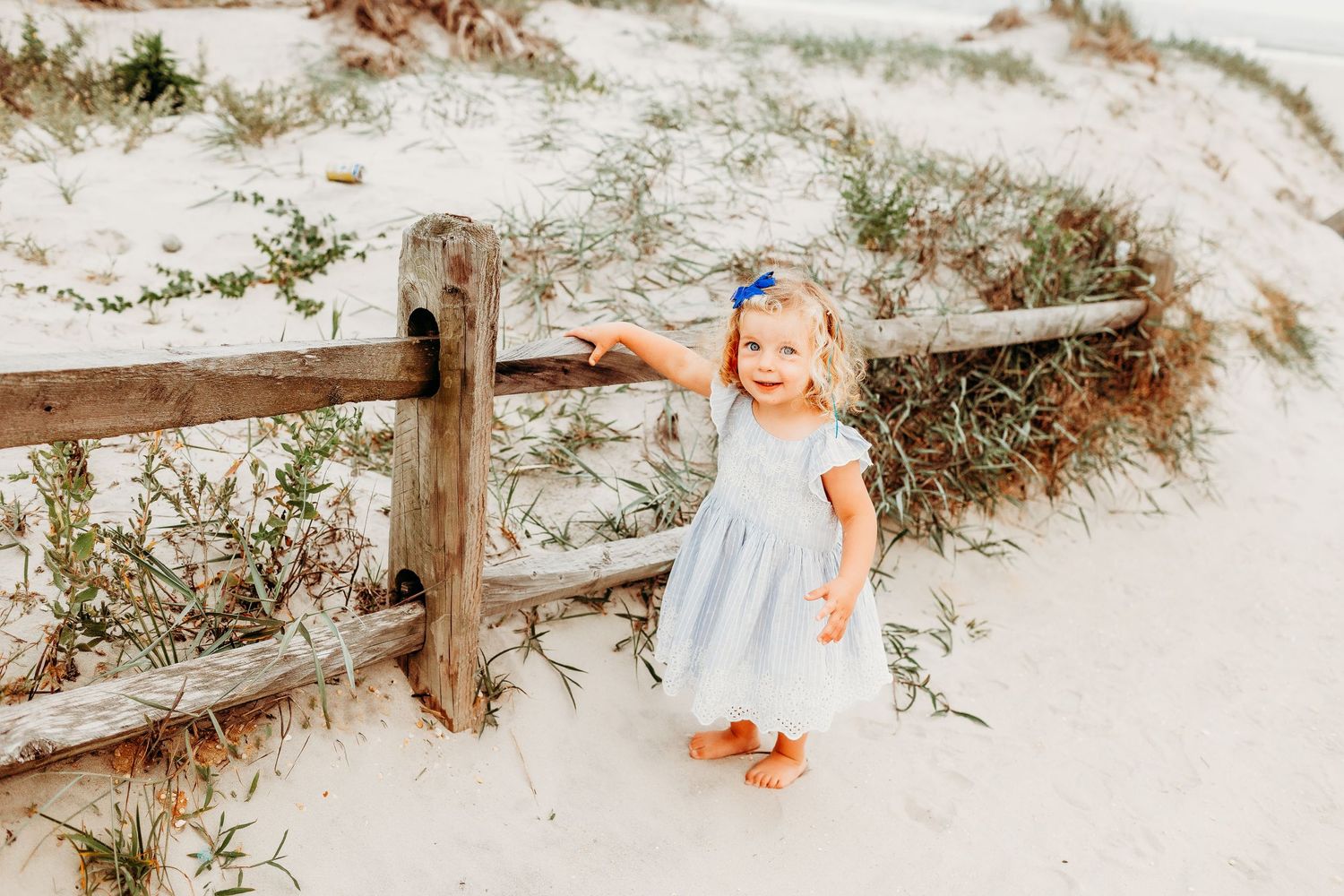 Child in a blue dress stands by a wooden fence on a sandy beach path.