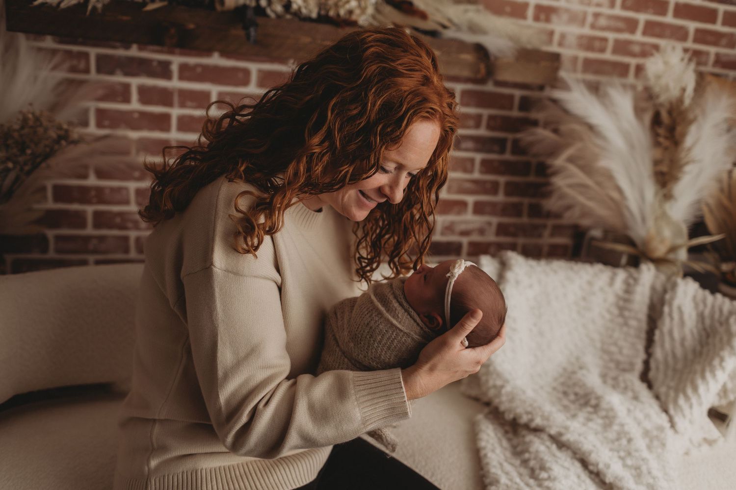 Red-haired mother in cream sweater smiling while cradling her newborn baby in a cozy studio setting in Ridley Park, pa