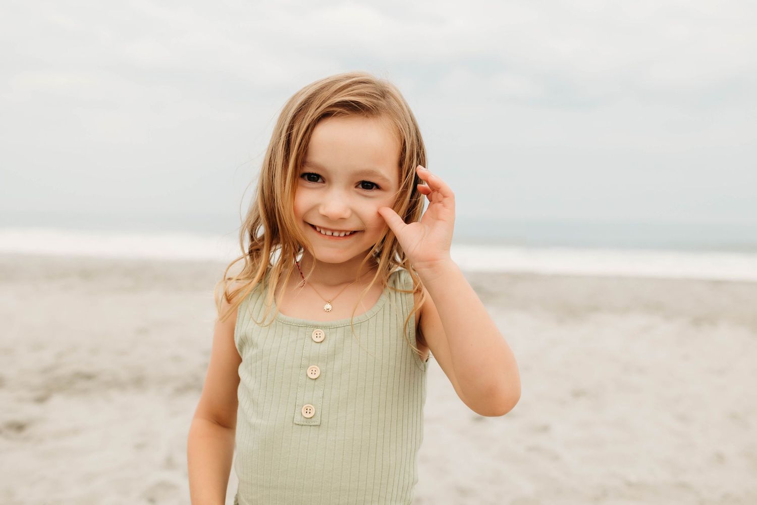 Young child wearing a mint green tank top smiles while standing on a cloudy beach.