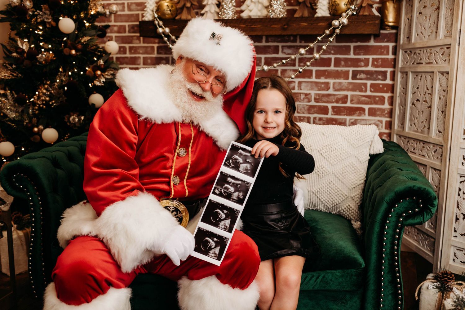 A person in a Santa suit sits on a green velvet chair with a child holding ultrasound photos during a holiday photo session.