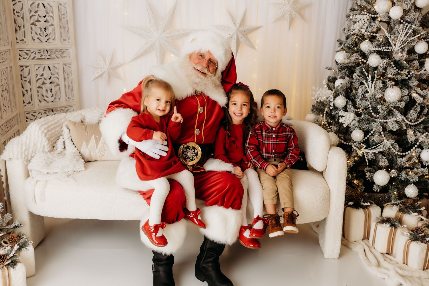 Three young children sit with Santa Claus on a white sofa beside a beautifully decorated Christmas tree.