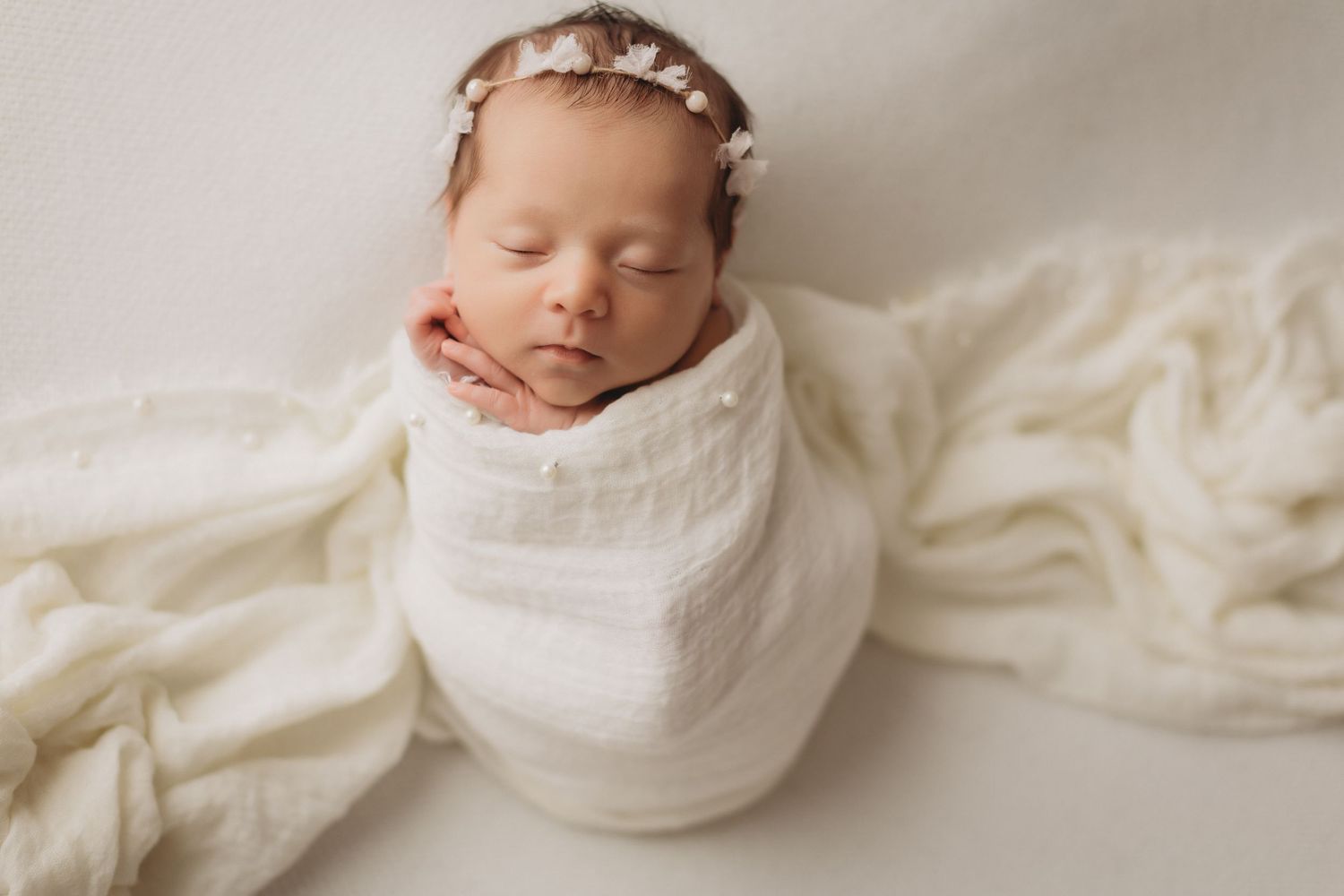 Peaceful newborn baby girl swaddled in white wrap with floral headband, sleeping on soft cream blanket.