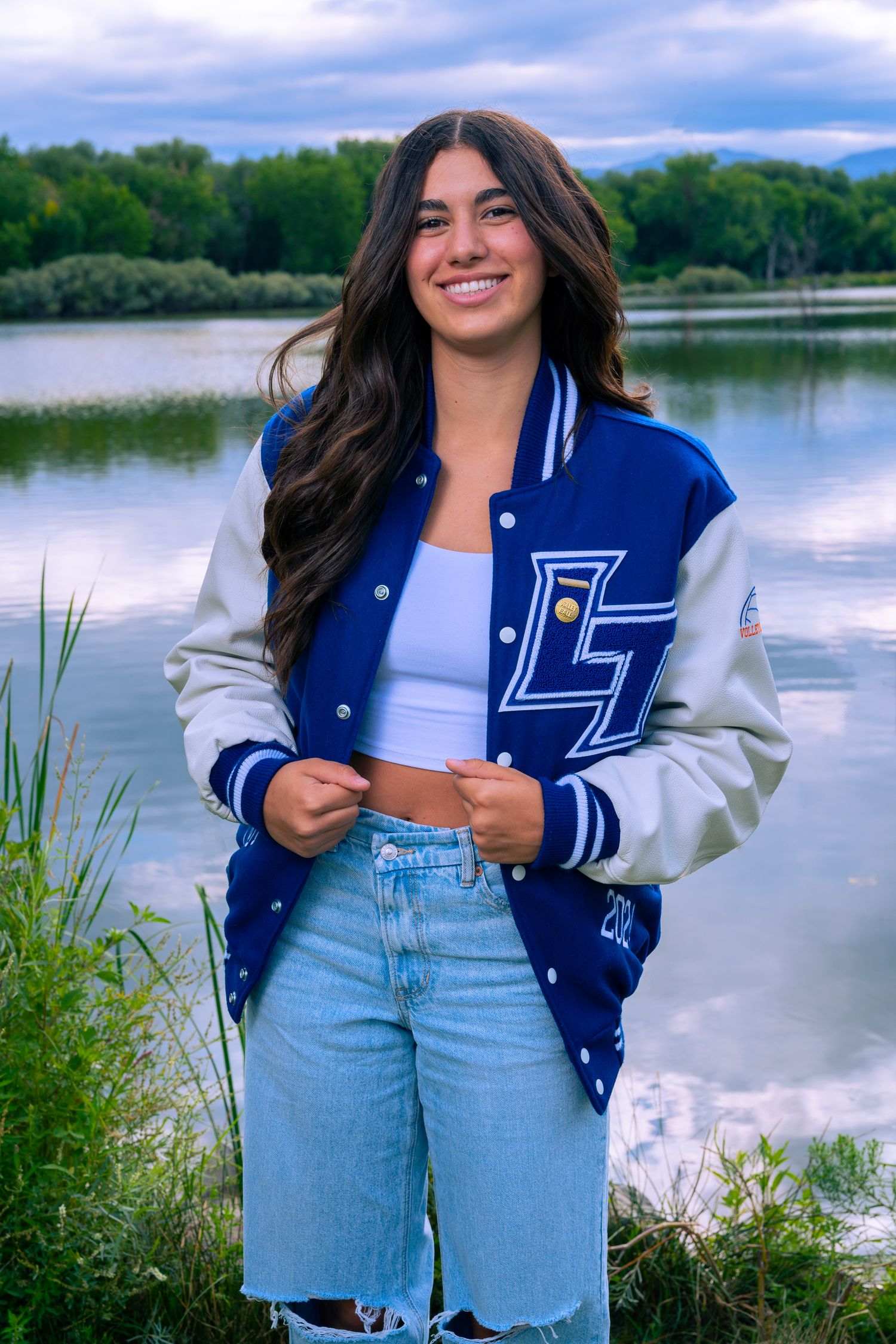 High school senior girl smiling near a lake wearing a letterman jacket in Northern Colorado