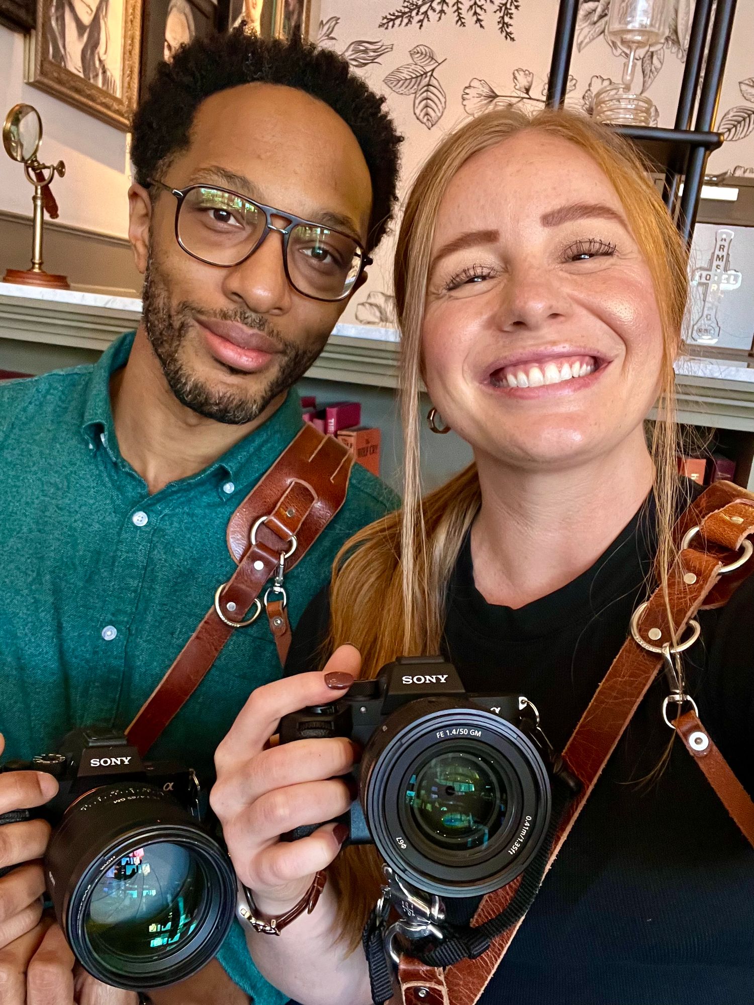 Close-up selfie of husband and wife dual-photographer team holding their cameras indoors in Northern Colorado