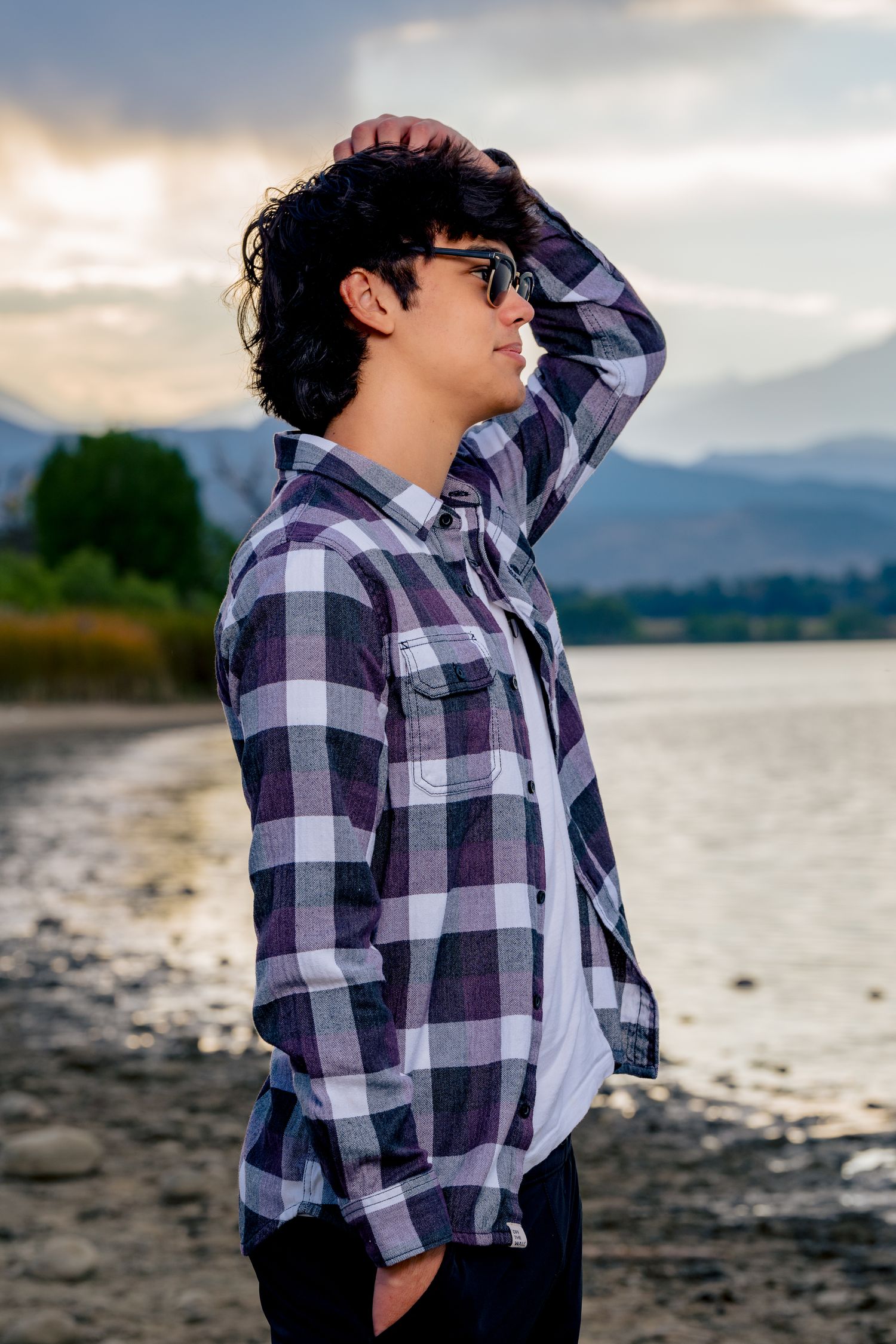 High school senior boy wearing sunglasses and a plaid shirt posing near a lake with mountain views in Northern Colorado