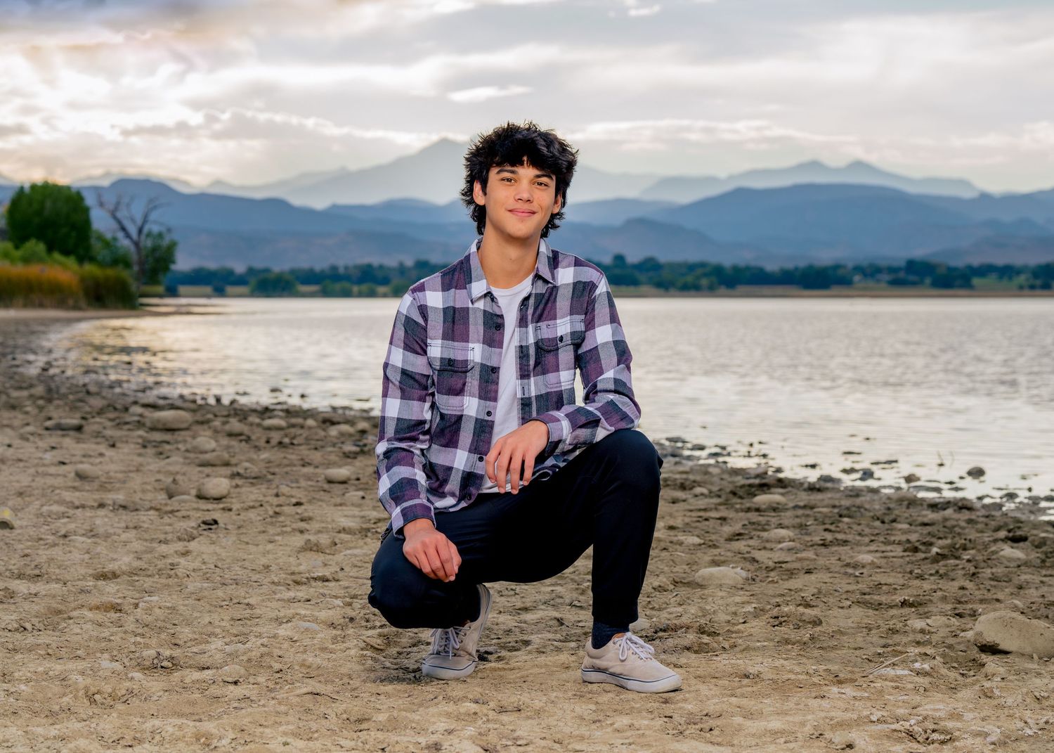 High school senior boy crouching on a sandy shore in front of mountain foothills in Northern Colorado