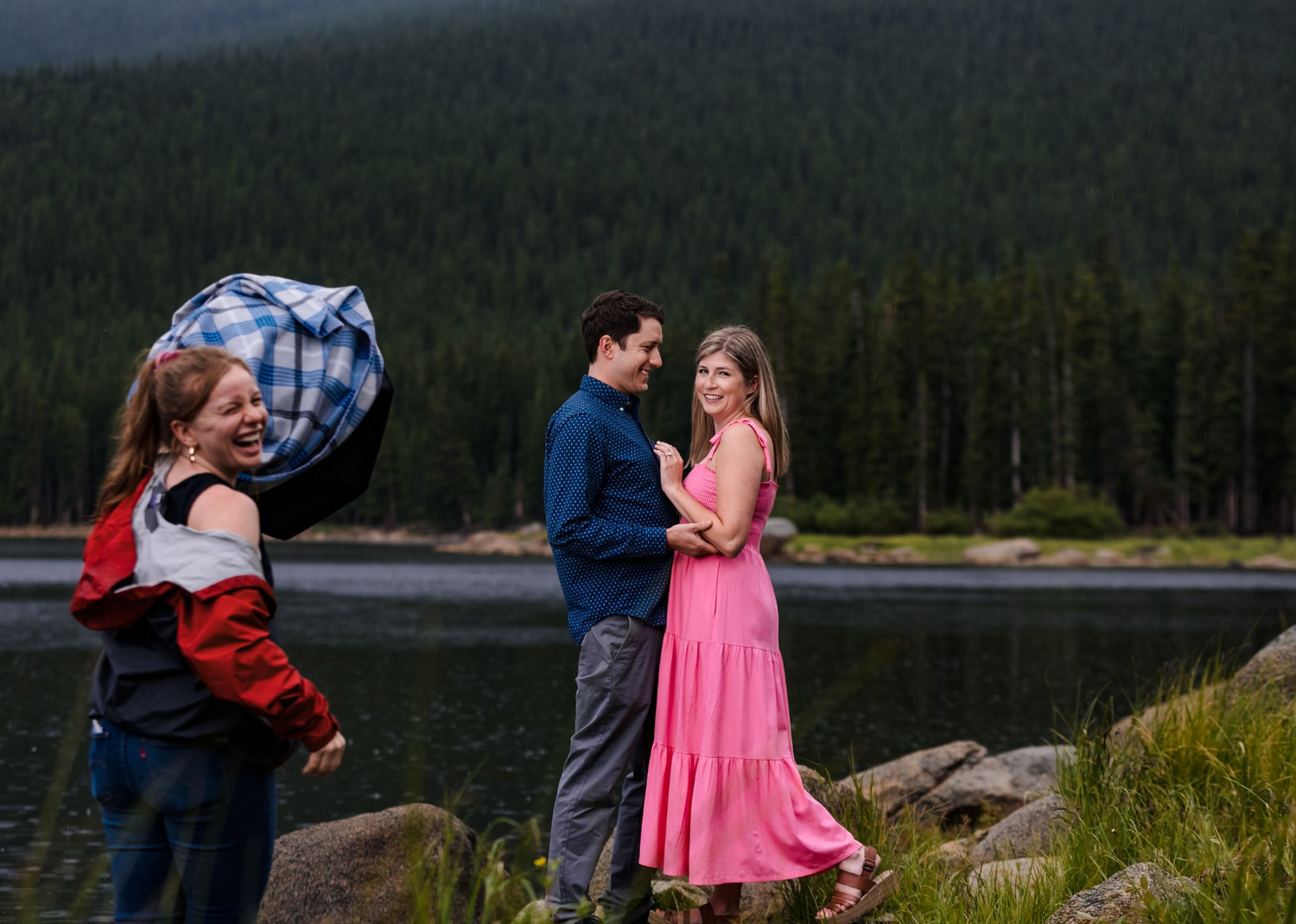Behind the scenes with female photographer holding lighting diffuser while couple poses by a mountain lake in Northern Colorado