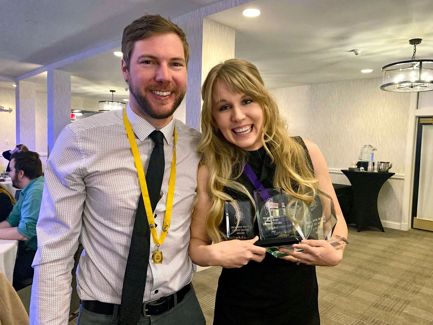 Smiling man in a white shirt and gold medal lanyard stands beside a blonde woman holding a glass award at an indoor event.