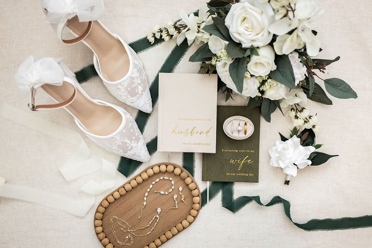 Flat lay of lace wedding heels, white floral bouquet, vow booklets, greenery ribbon and a beaded wooden tray on a cream background.
