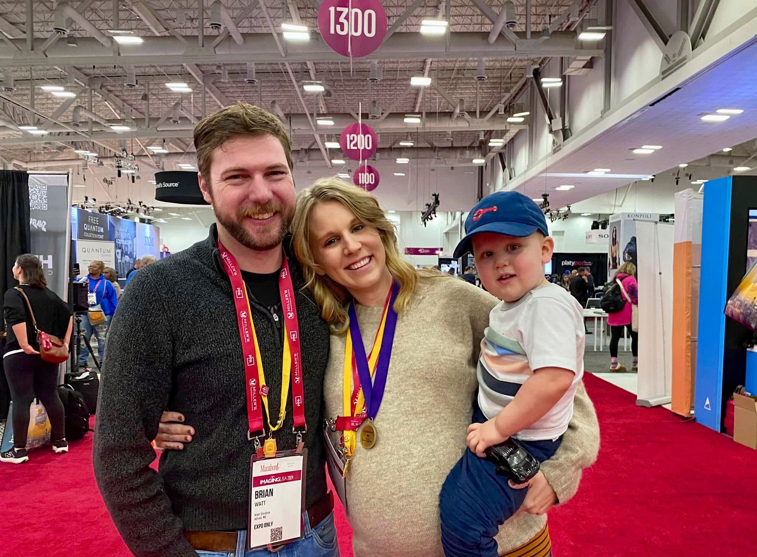 A smiling man and woman wearing lanyards and medals hold a toddler in a blue cap at a busy convention hall with red carpet.