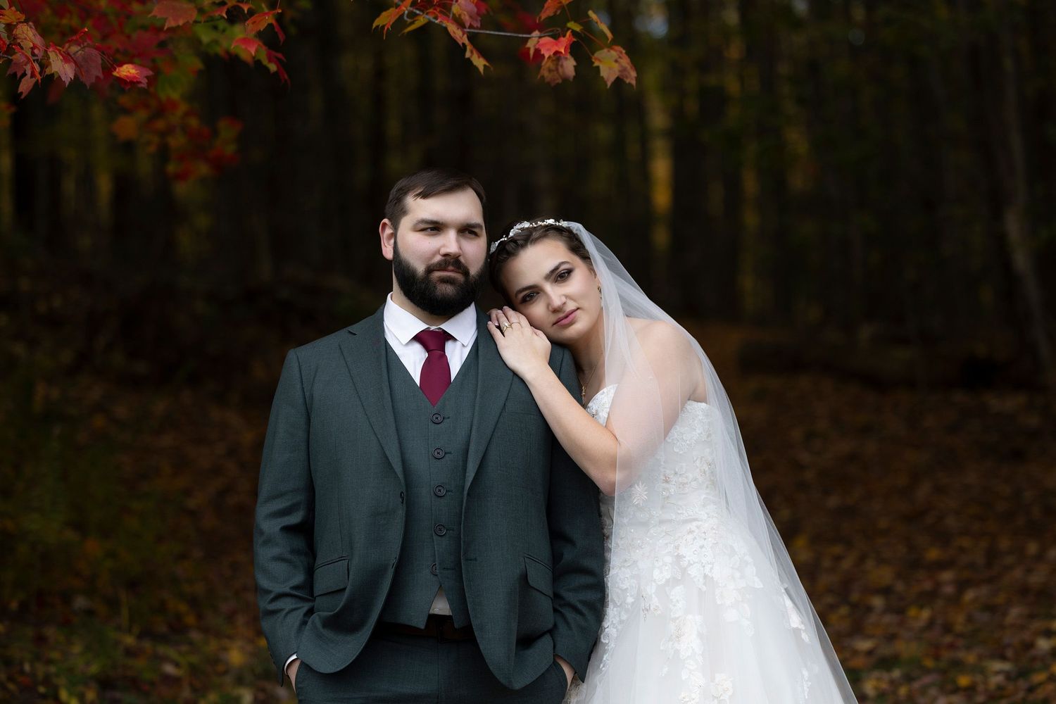 Bride in a white lace gown rests her head on the shoulder of a groom in a gray suit and burgundy tie in an autumn forest.
