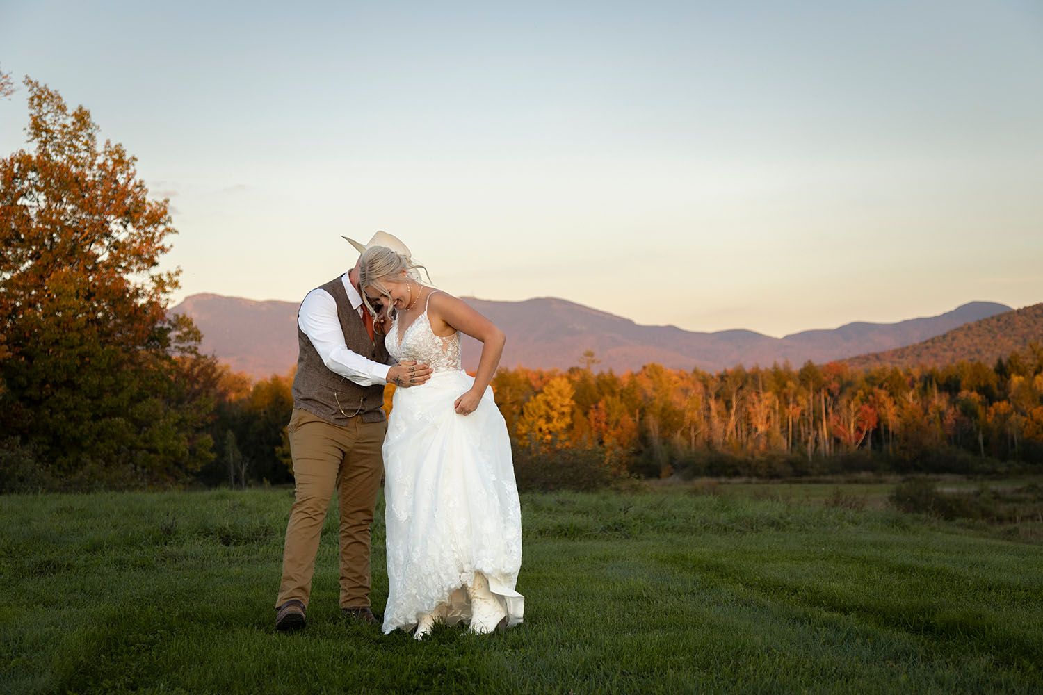 Bride in a white gown and groom in a vest and hat share a kiss on a grassy field with autumn foliage and mountains at sunset.