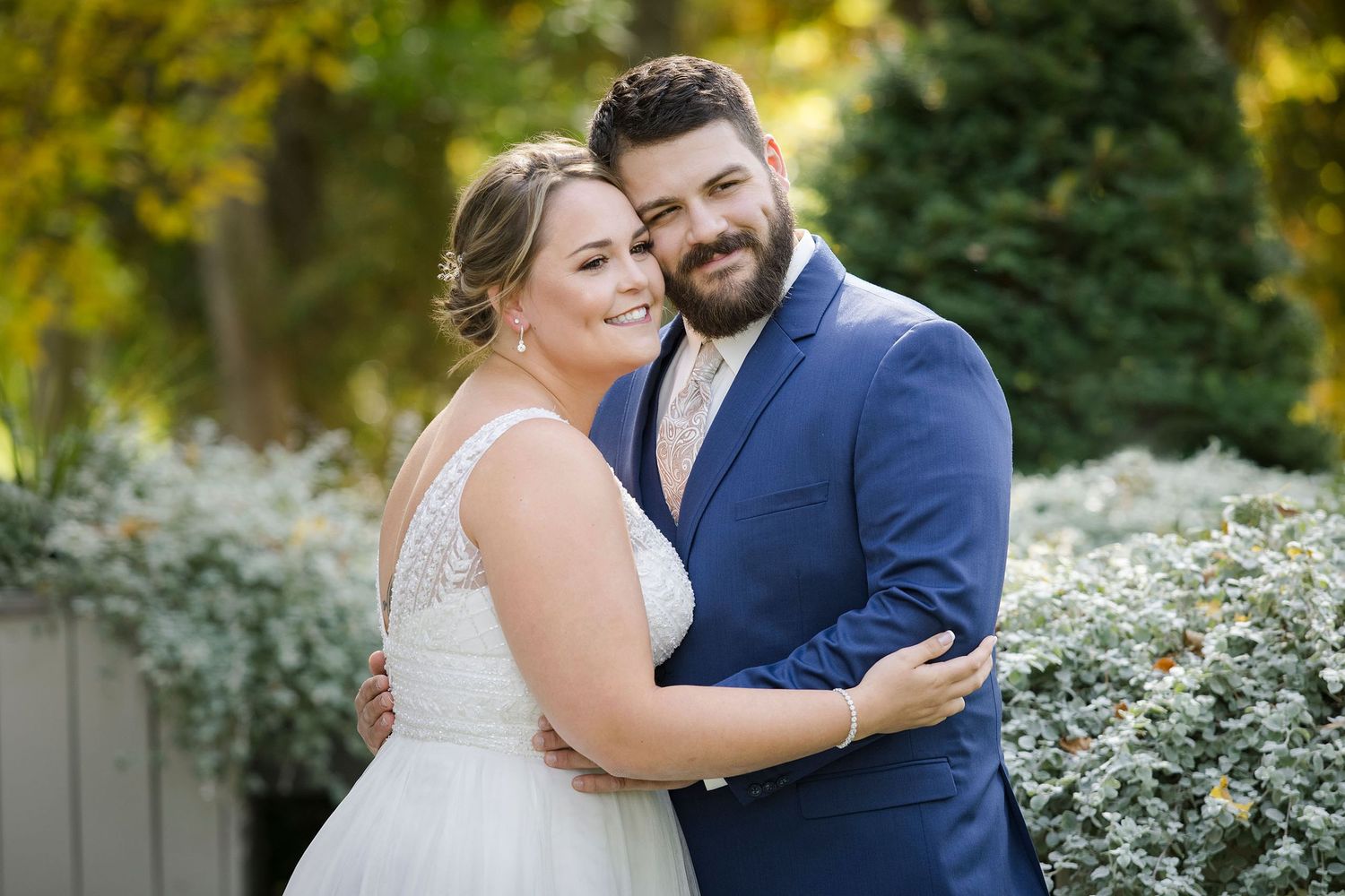 Bride and groom embrace outdoors in New England garden setting, she in white gown, he in blue suit.