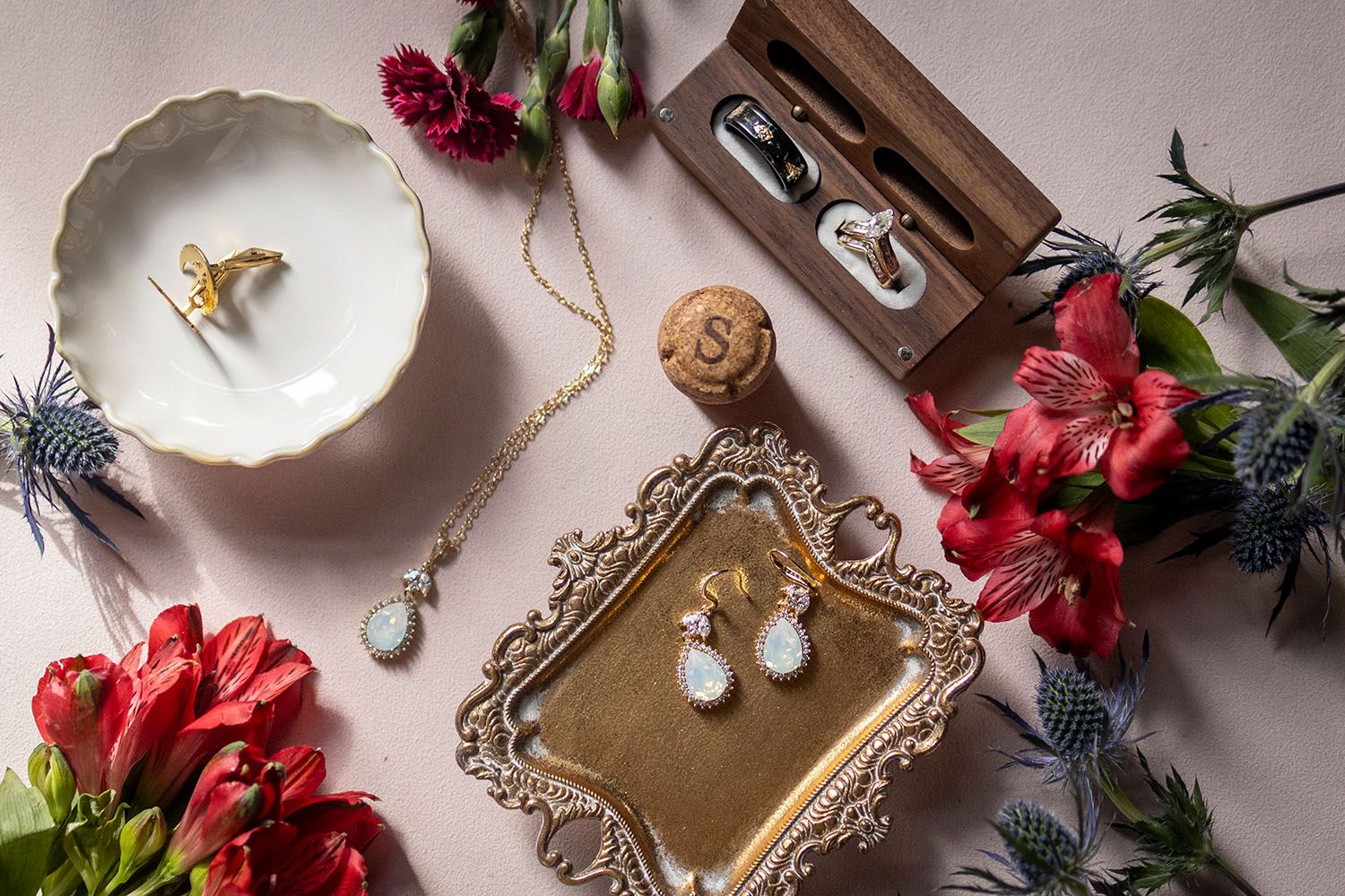 Pearl drop earrings, a pendant necklace and wedding rings arranged with red alfreda flowers and blue thistles on a blush pink backdrop.