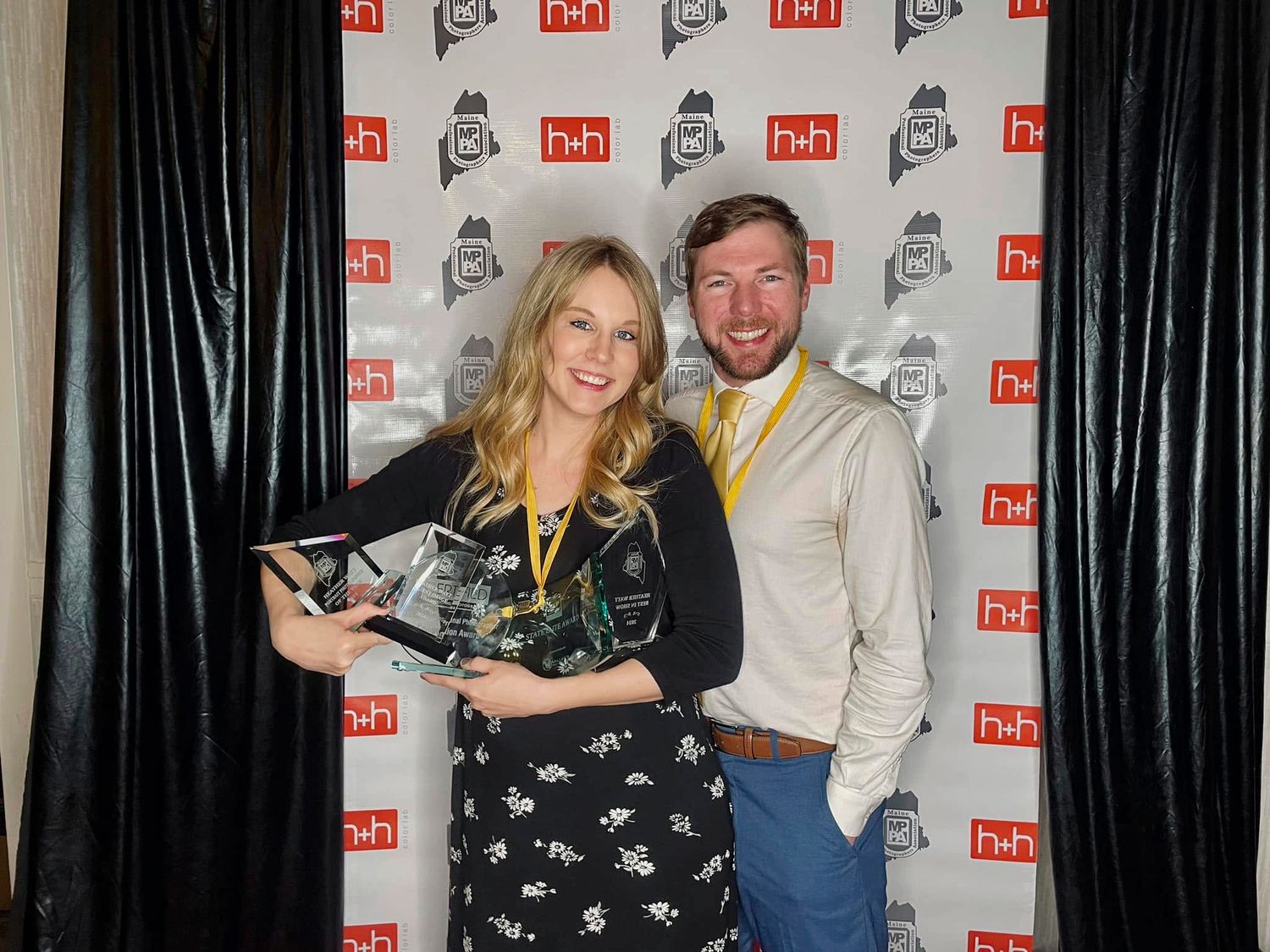 Smiling woman in a black floral dress holds a glass award beside a bearded man in a tan shirt at a step-and-repeat banner event.