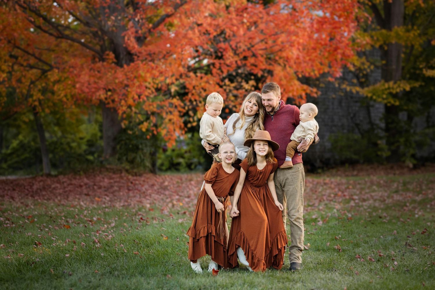Family of six posing in park with vibrant red and orange fall foliage backdrop.