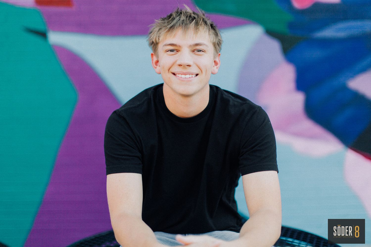 high school senior boy sitting and smiling in front of colorful mural wall in charles city, iowa - SODER 8