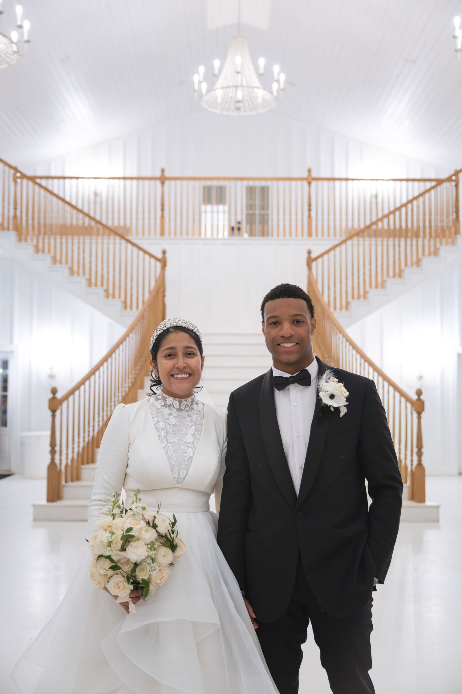 Bride and groom posing in elegant white venue with grand staircase and chandelier overhead. Ivy Rose Barn, Rocky Mount, VA