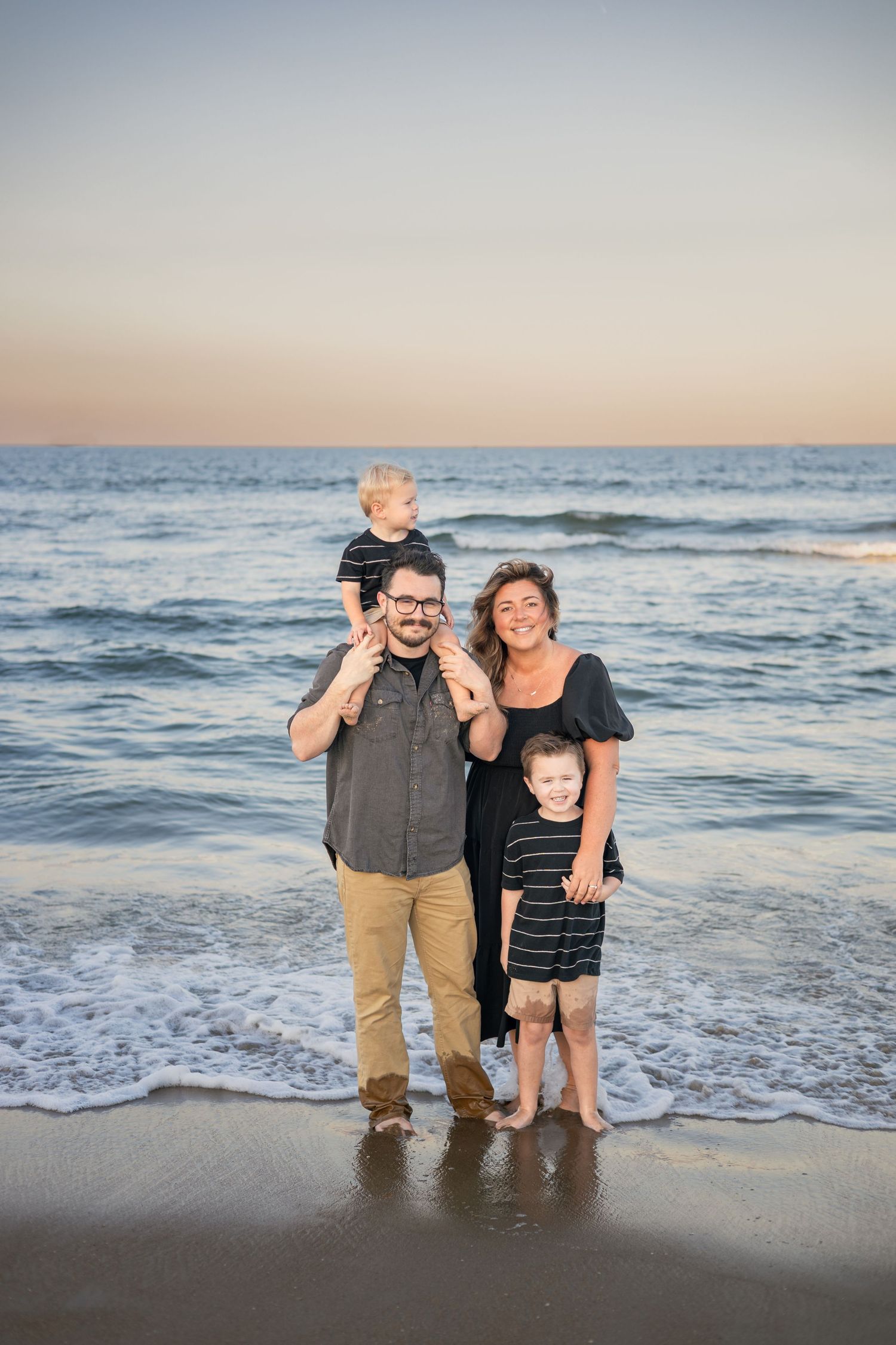 Family of four posing on a beach at sunset, two parents with two young boys, waves gently lapping at their feet.