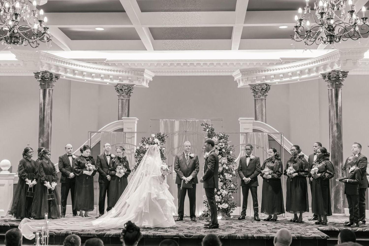 Black and white wedding ceremony photo with bride, groom, and wedding party standing at elegant venue altar.