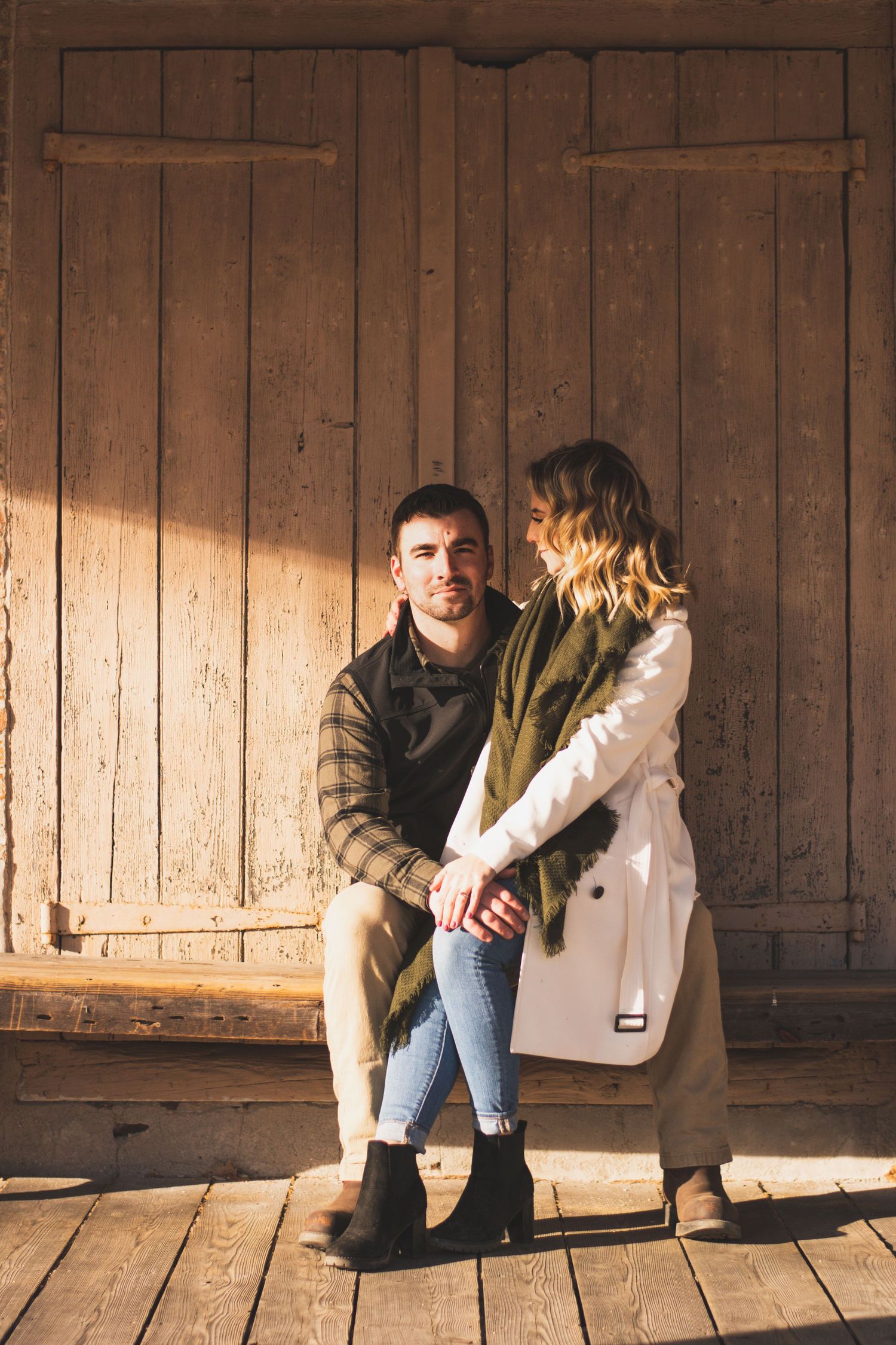 Couple sitting together holding hands on a sunlit wooden porch against a rustic barn door.
