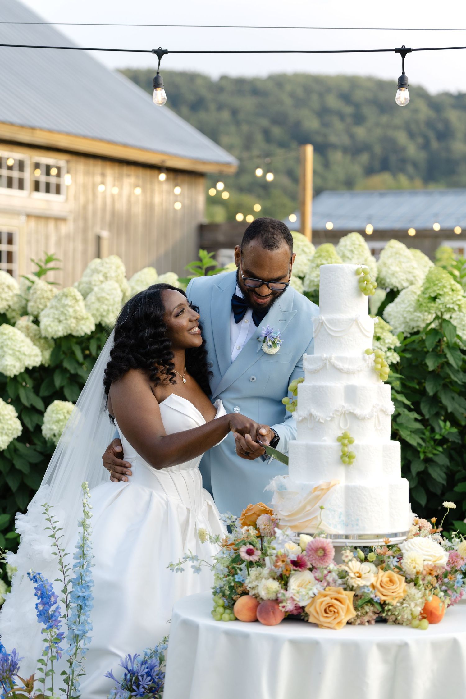 Couple cutting a tall white tiered wedding cake outdoors surrounded by hydrangeas and colorful blooms.
