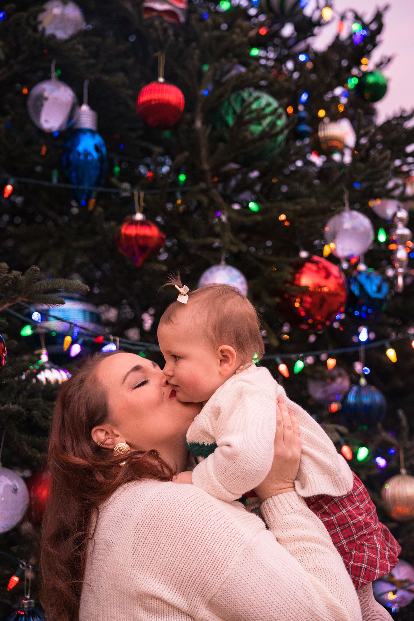 Mother kissing baby girl in front of a decorated Christmas tree with colorful ornaments and twinkling lights.
