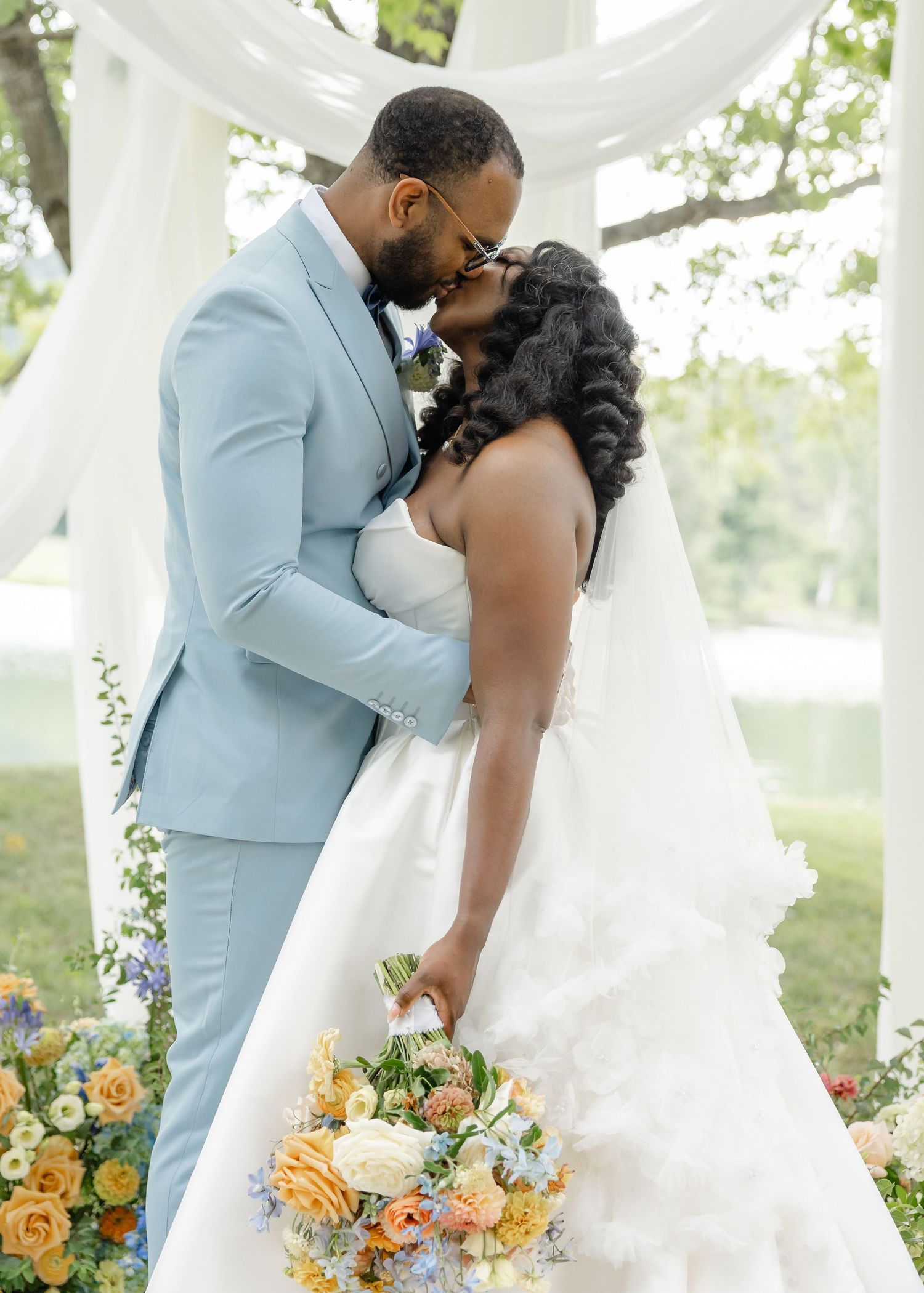 African American couple sharing a kiss under floral arch at outdoor garden wedding ceremony.