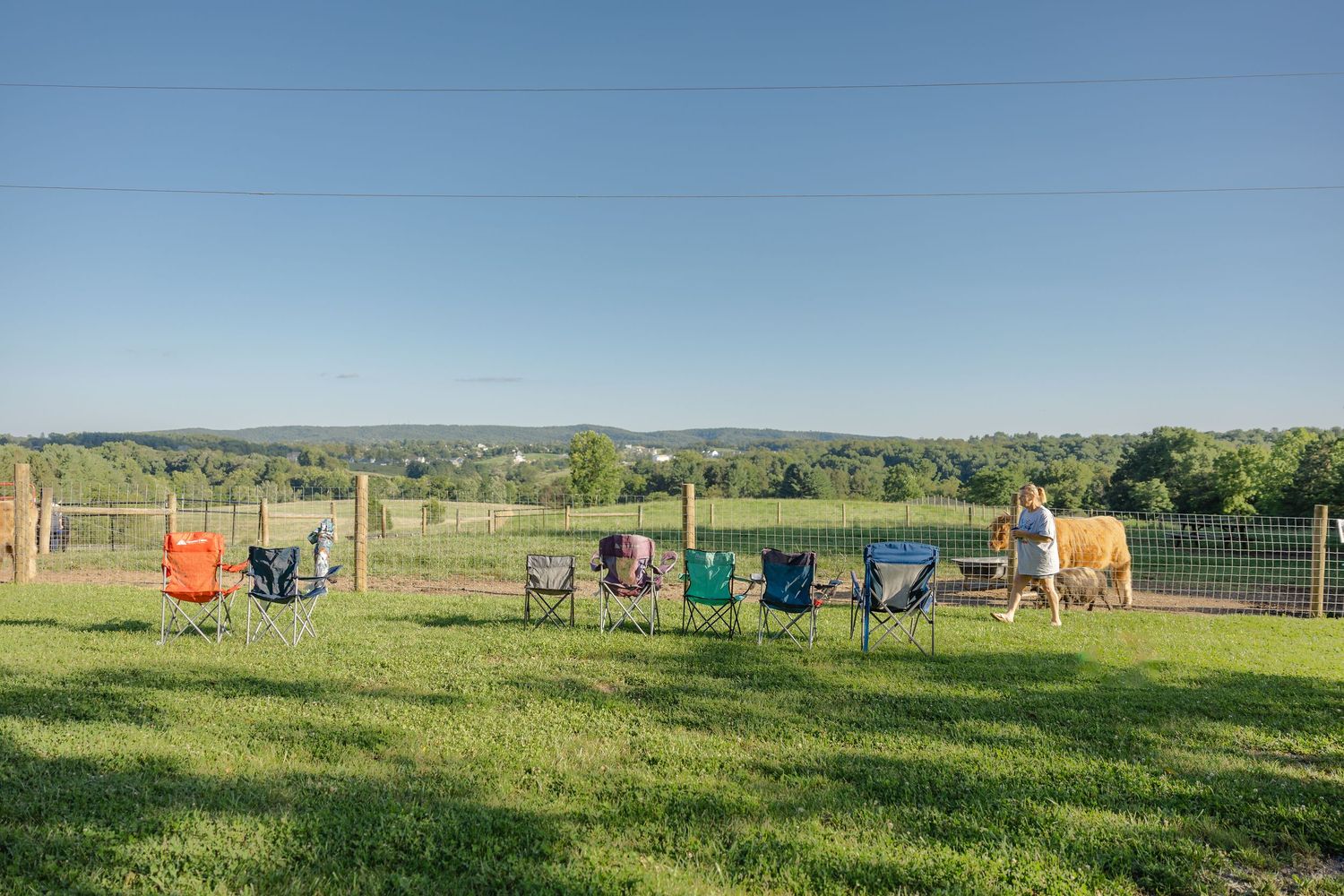 Colorful folding chairs arranged in a row on green grass with a scenic countryside view and fence in background.