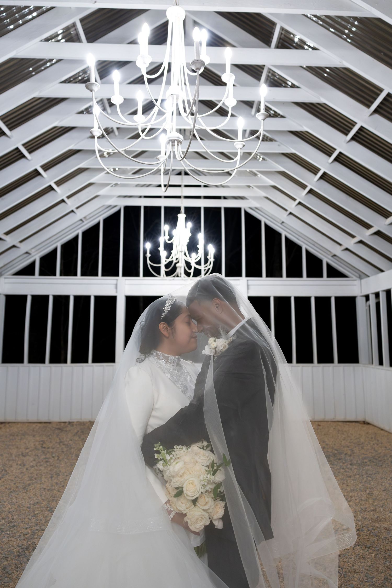 Bride and groom kiss under wedding veil in white chandelier gazebo venue.