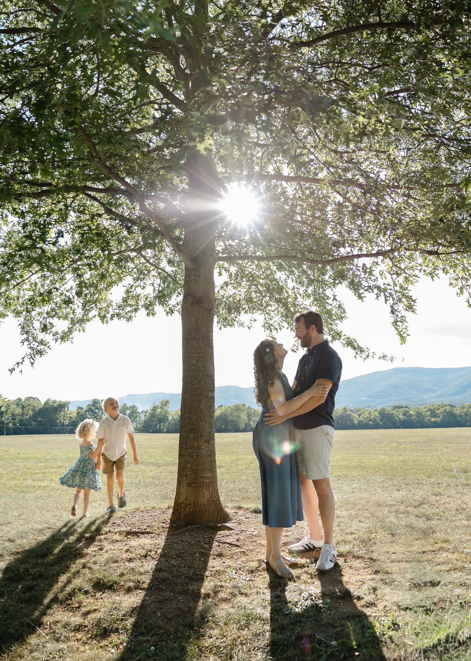 Couple embracing under a sunlit tree while two children run through an open field with mountains.