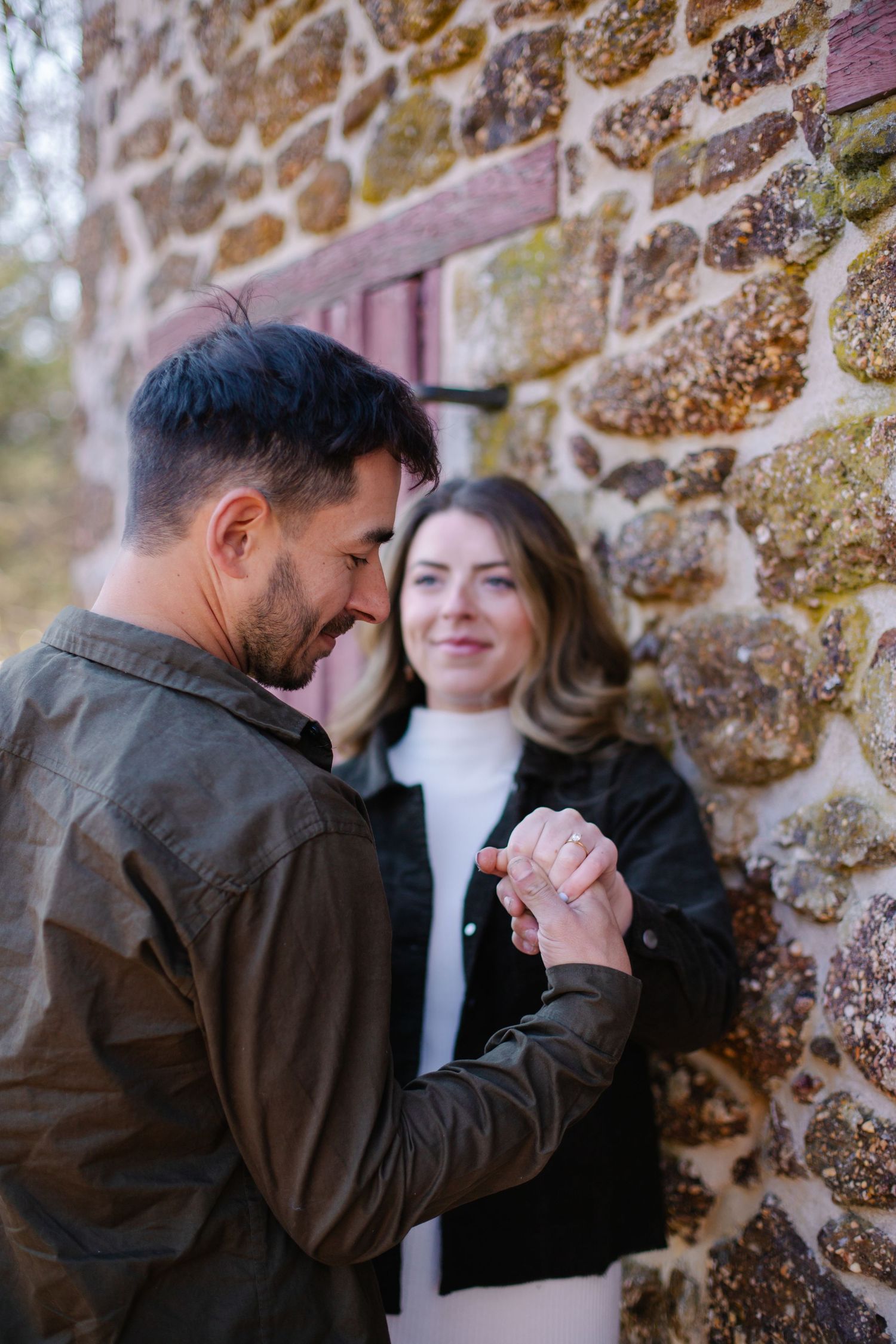 Man in olive jacket admiring engagement ring on smiling womans finger by a stone wall.