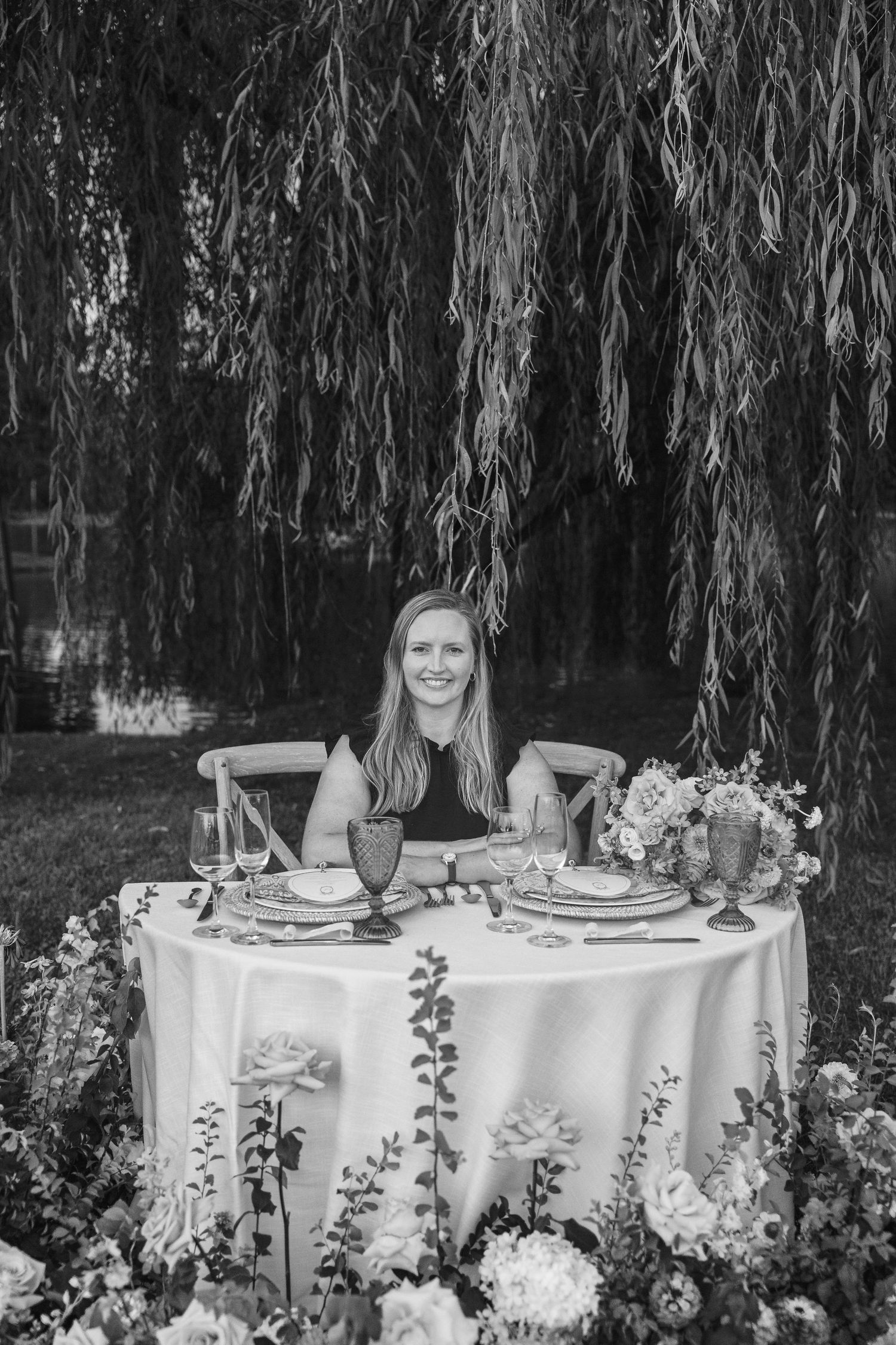 Woman smiling at elegantly set outdoor table beneath weeping willow tree, surrounded by floral arrangements, black and white photo.